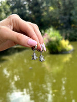 Hand holding a pair of silver frog-shaped earrings with a blurred natural background