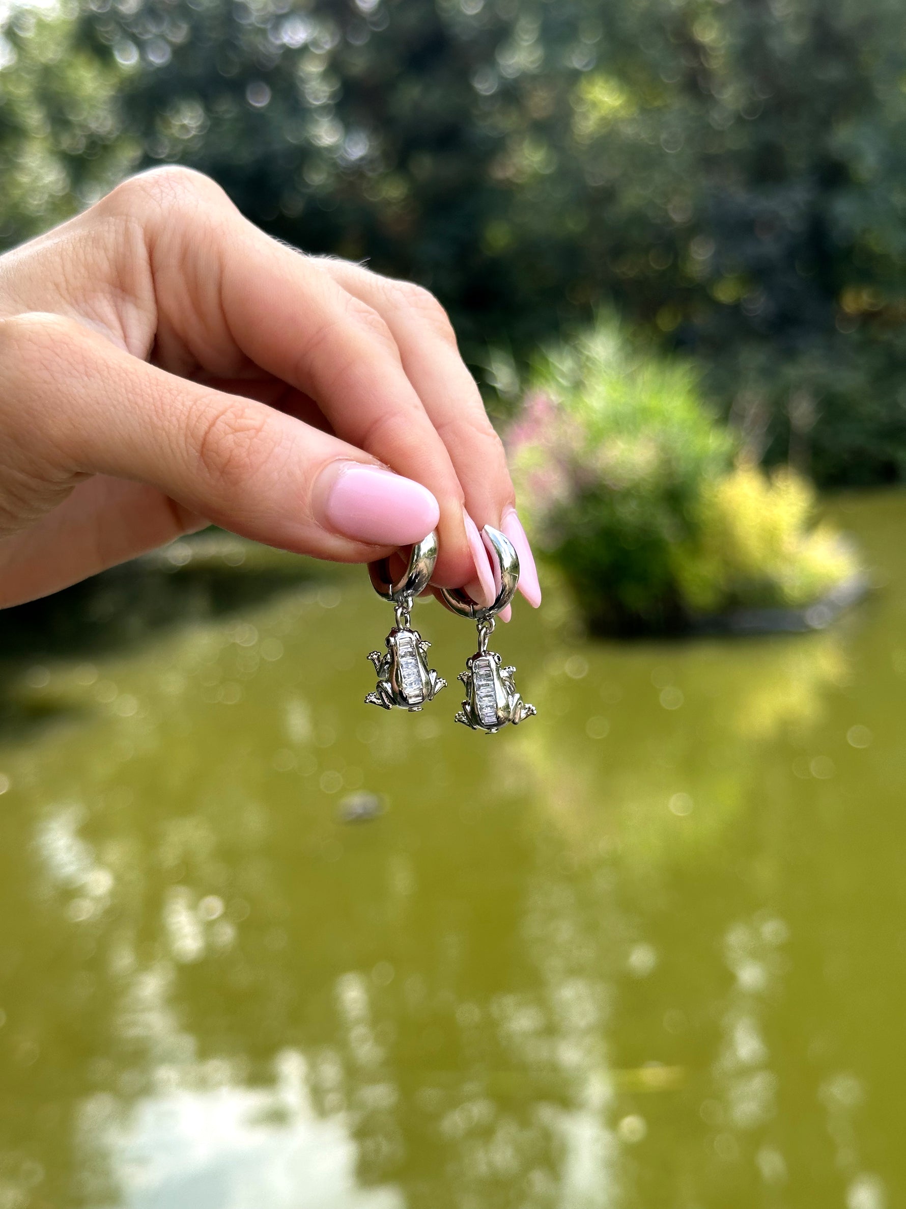 Hand holding a pair of silver frog-shaped earrings with a blurred natural background
